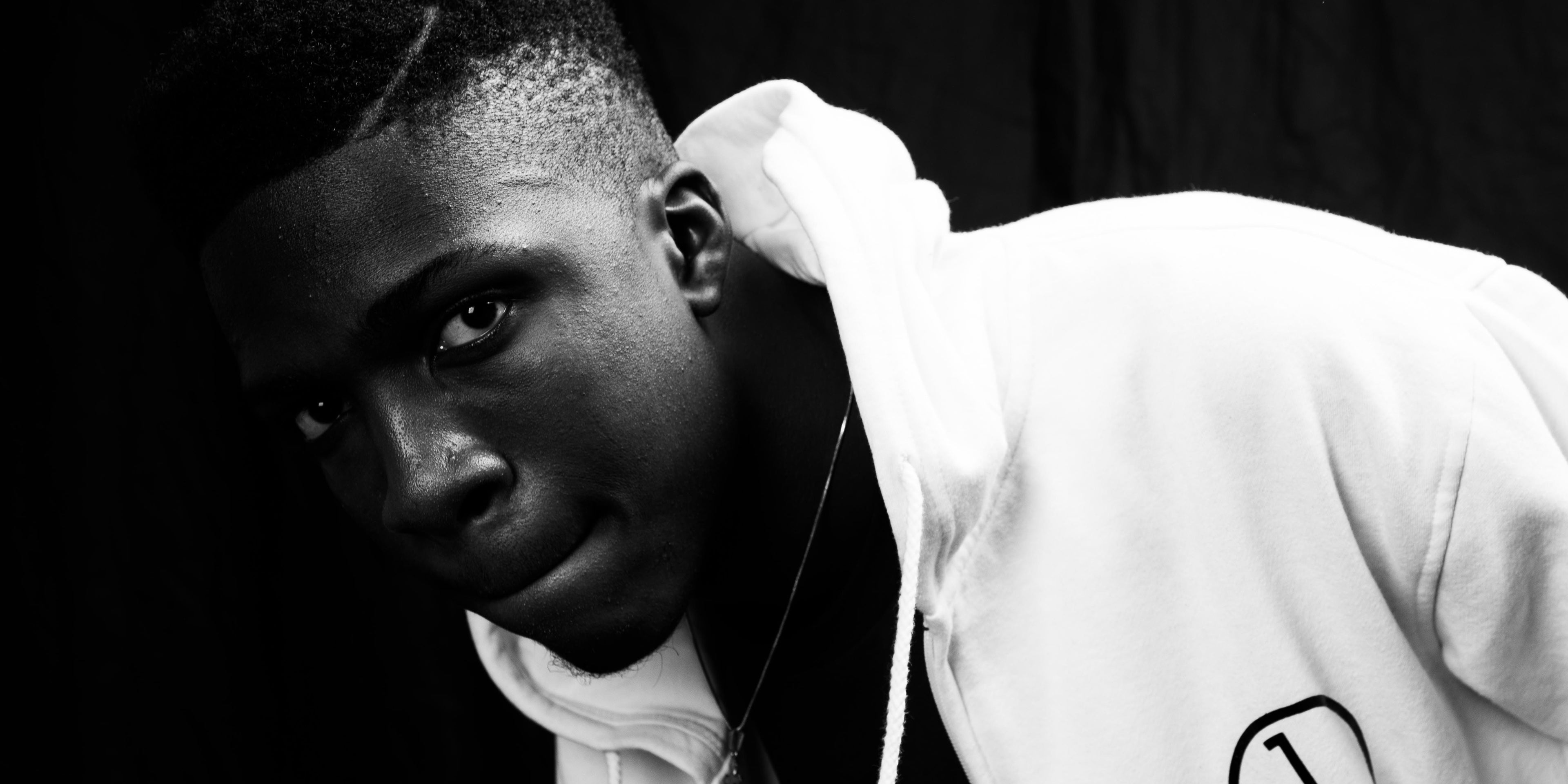 Close portrait leaning into frame, wearing a white zip hoodie with the house mark, against a black ground